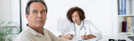 Patient mittleren Alters mit Diabetes sitzt neben verschwommenem afrikanisch-amerikanischem Arzt in Klinik, Banner 