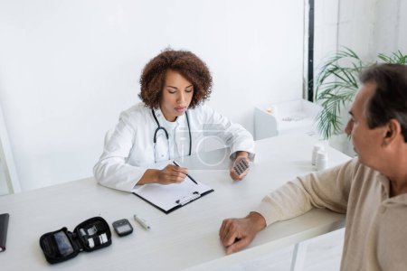 Afrikanisch-amerikanischer Arzt hält Tabletten in der Hand und blickt auf Klemmbrett in der Nähe eines Patienten mit Diabetes in der Klinik 