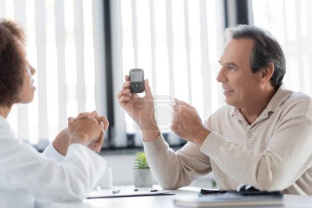 Positive mature patient holding glucometer near blurred african american doctor in clinic 