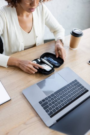 Vue recadrée d'une femme d'affaires afro-américaine tenant un kit de diabète près d'un ordinateur portable et d'un café au bureau 