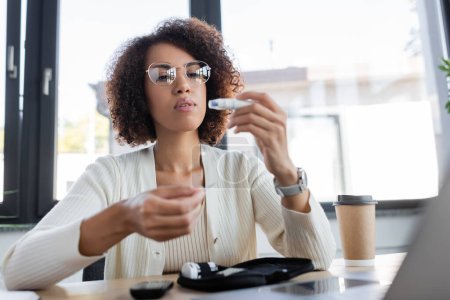 Afroamerikanische Geschäftsfrau blickt im Büro auf Glukometer-Lanzette neben Laptop 
