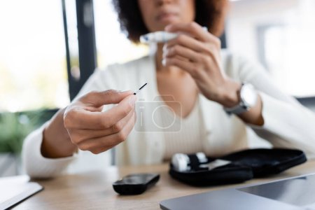 Ausgeschnittene Ansicht einer verschwommenen Afroamerikanerin mit Teststreifen des Glukometers im Büro 
