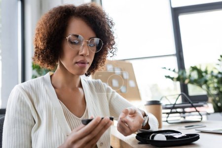 Afroamerikanische Geschäftsfrau mit Brille kontrolliert Blutzucker mit Glukometer im Büro 