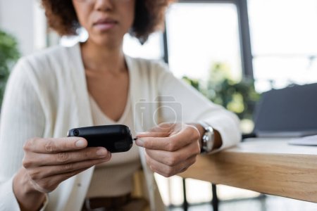 Cropped view of blurred african american businesswoman using glucometer while working in office 
