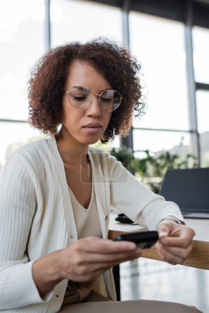 Téléchargez les photos : Femme d'affaires afro-américaine en lunettes regardant le glucomètre flou au bureau - en image libre de droit