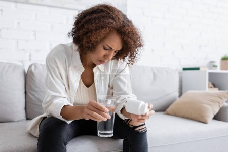 African american woman with diabetes feeling bad while holding pills and water on couch at home 