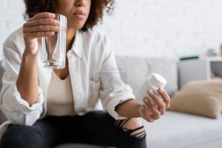 Vue recadrée d'une femme afro-américaine diabétique tenant un pot avec des pilules et un verre d'eau à la maison 