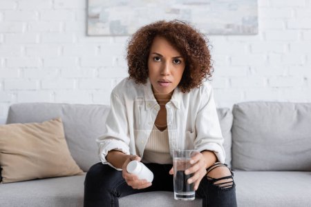 Mujer afroamericana con diabetes sosteniendo pastillas y agua mientras mira la cámara en casa 