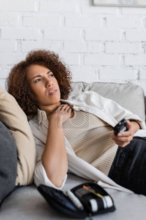 Afroamerikanerin mit Diabetes fühlt sich unwohl und hält Glukometer in der Nähe medizinischer Ausrüstung auf der Couch 
