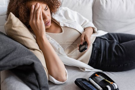 Afroamerikanerin mit Diabetes berührt Kopf und hält Glukometer auf Couch 