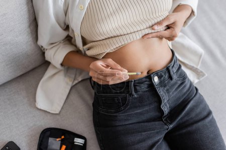 Vista superior de una mujer afroamericana inyectándose insulina en el vientre cerca del kit de diabetes en casa 