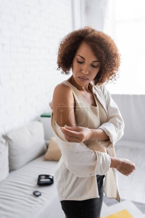 Mujer afroamericana joven con diabetes inyectándose insulina en el brazo en la sala de estar 