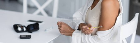 Ausgeschnittene Ansicht einer afrikanisch-amerikanischen Frau bei der Insulinspritze in der Nähe eines verschwommenen Glukometers in der Küche, Banner 