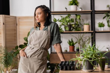 young african american florist standing with hand in pocket of apron and looking away in flower shop