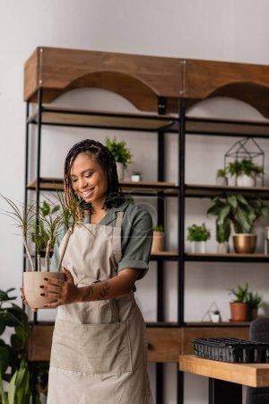 alegre florista afroamericana en delantal sosteniendo planta en maceta verde cerca de estante borroso en tienda de flores