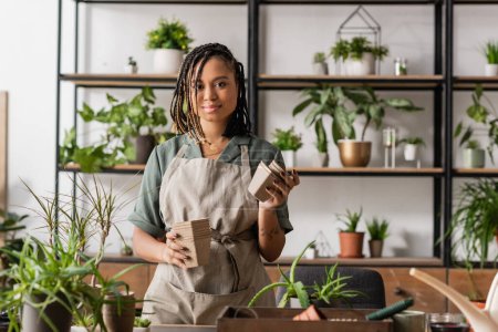 young african american florist holding peat pots near green plants and blurred rack in flower shop