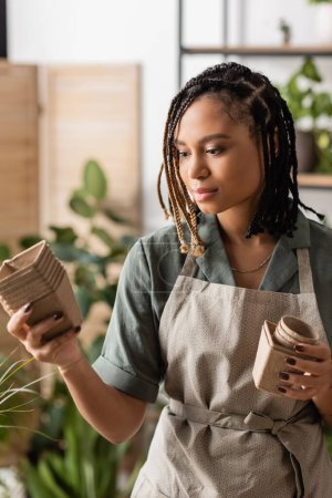 Téléchargez les photos : Jeune fleuriste afro-américain avec des tresses tenant des pots de tourbe dans un magasin de fleurs - en image libre de droit