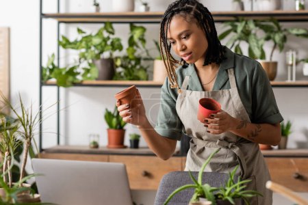 stylish african american woman showing flowerpots during video call on laptop in flower shop