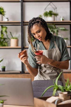 young african american woman in apron holding flowerpots near laptop during video call in flower shop