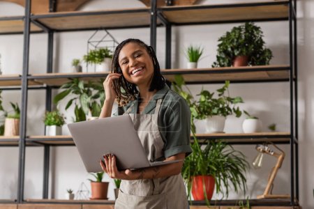 cheerful african american florist with dreadlocks standing with laptop near rack with green potted plants on blurred background