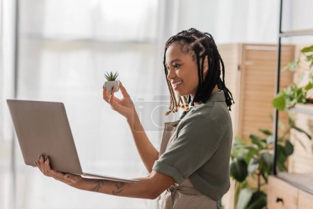 happy african american florist with dreadlocks showing small potted plant during video call in flower shop