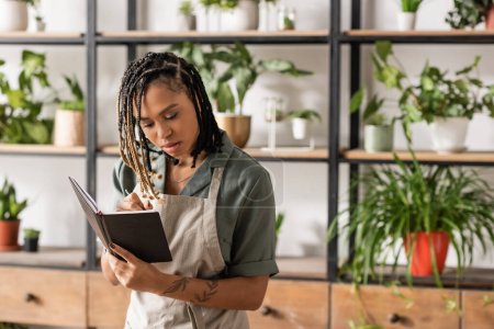 young african american florist in apron writing order in notebook in flower shop