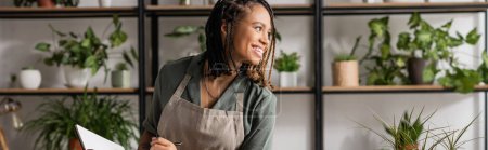 cheerful african american florist holding notebook and looking away near rack with plants on blurred background, banner
