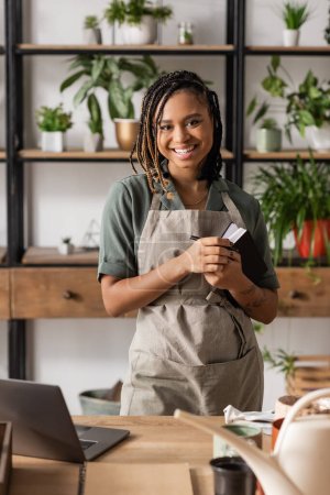 trendy african american woman with copybook looking at camera near laptop and potted plants on blurred background