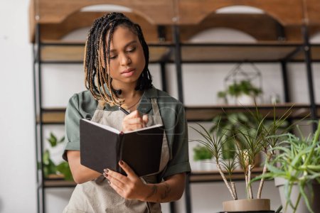young african american florist with braids writing order in notebook near green plants in flower shop