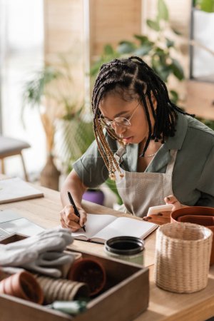 stylish african american florist in apron and eyeglasses writing order in notebook while sitting with smartphone in flower shop