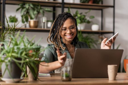 heureux fleuriste afro-américain tenant smartphone et geste lors d'un appel vidéo sur ordinateur portable près de plantes floues