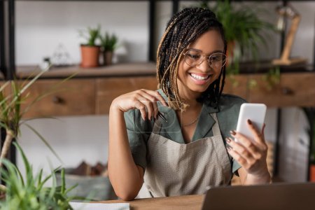 Téléchargez les photos : Joyeux fleuriste afro-américain avec coiffure à la mode et lunettes regardant téléphone portable dans le magasin de fleurs - en image libre de droit