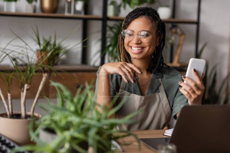 happy african american florist in eyeglasses holding smartphone and looking at green plants on blurred foreground