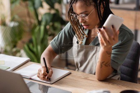 junge afrikanisch-amerikanische Floristin mit Brille hält Smartphone in der Hand und schreibt in Notizbuch neben verschwommenem Laptop in Blumenladen