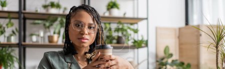 Photo for African american florist in stylish eyeglasses and dreadlocks holding coffee to go and looking at camera in flower shop, banner - Royalty Free Image