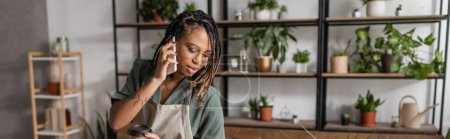 young african american florist in eyeglasses talking on mobile phone near potted plants on blurred background, banner