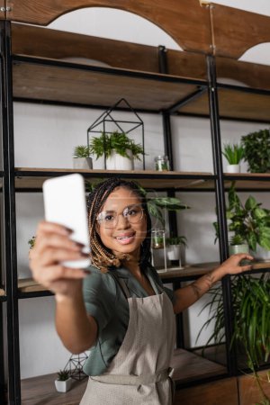 Foto de Floristería afroamericana joven y elegante sosteniendo teléfono inteligente borroso mientras que muestra rack con surtido de plantas durante la videollamada - Imagen libre de derechos