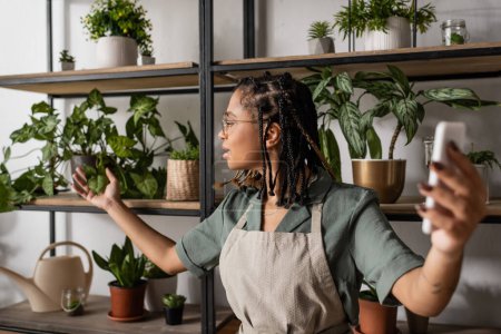 african american florist in apron holding smartphone and touching leaves of green plant during online trade