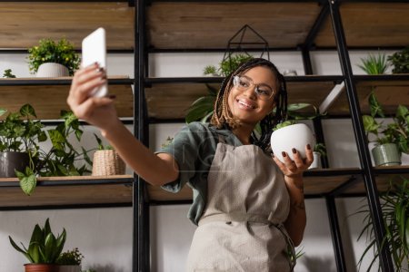 Foto de Floristería afroamericana feliz sosteniendo la planta en maceta verde y el teléfono celular durante el pedido en línea en la tienda de flores - Imagen libre de derechos