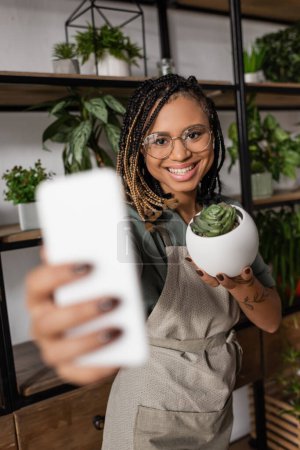 Foto de Alegre florista afroamericano sosteniendo teléfono móvil borroso y planta en maceta verde durante la videollamada en la tienda de flores - Imagen libre de derechos