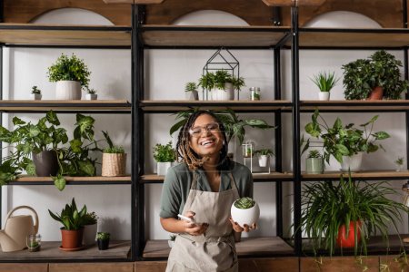 happy african american florist with mobile phone looking at camera near rack with different potted plants
