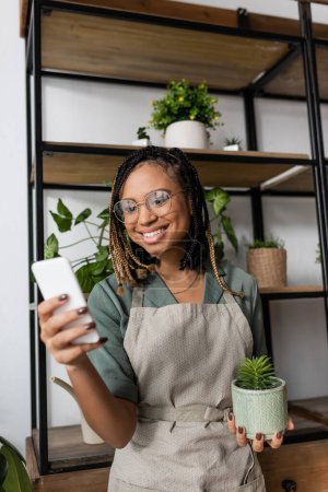 Foto de Floristería afroamericana feliz sosteniendo la planta en maceta y el teléfono móvil durante la videollamada en la tienda de flores - Imagen libre de derechos