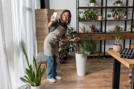 Photo for Happy african american florist holding smartphone and showing potted tropical plant in flower shop - Royalty Free Image
