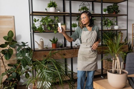 souriant afro-américain fleuriste dans tablier tenant téléphone portable tout en se tenant debout avec rack de plantes en pot lors de la commande en ligne