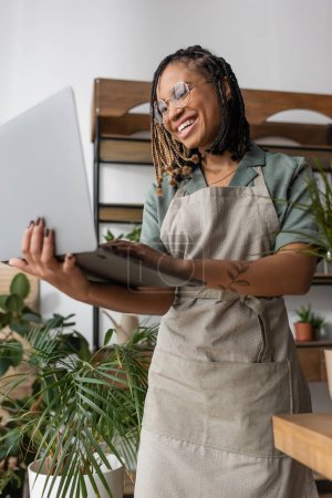 Low-Winkelansicht eines afrikanisch-amerikanischen Blumenhändlers mit Brille und Schürze, der mit Laptop im Blumenladen steht