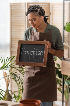 young and trendy african american florist in apron holding board with franchise lettering in flower shop