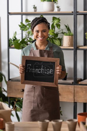 cheerful african american florist with trendy hairstyle holding board with franchise lettering near rack with plants