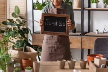 cropped view of african american florist in apron holding board with franchise lettering near plants and flowerpots on blurred foreground
