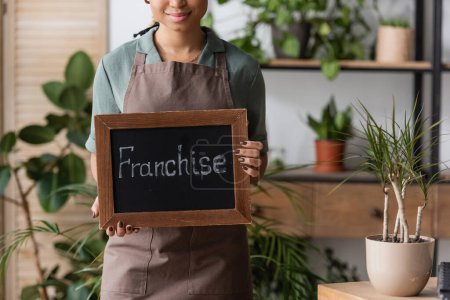 cropped view of african american florist in apron standing with board with franchise lettering near potted plant