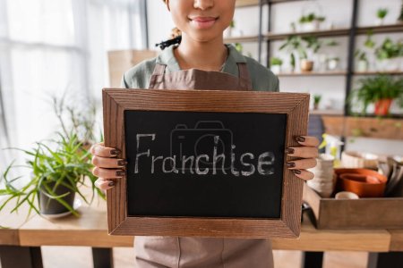 partial view of african american florist holding board with franchise lettering near plants and flowerpots on blurred background
